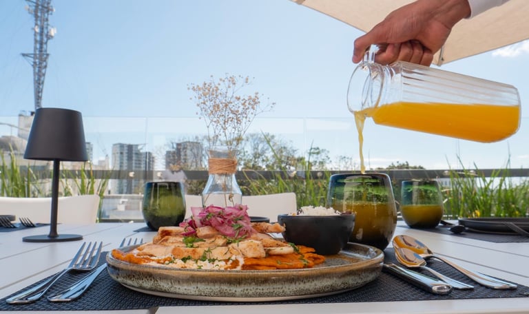 Hand pouring juice over a plated meal on a rooftop dining setting with city skyline in background