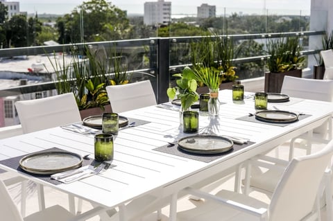 Outdoor rooftop dining table set with white chairs, place settings, and green potted plants overlooking a city skyline