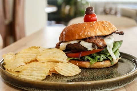 Gourmet burger with bacon and cheese on a seeded bun, topped with a cherry, served with crispy potato chips on a slate plate