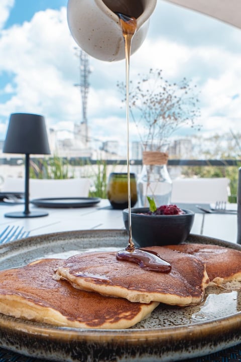 Golden pancakes on a plate with syrup being drizzled over them, viewed from a rooftop dining area with city skyline