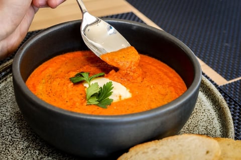 Hand drizzling sour cream into a bowl of bright orange soup garnished with fresh parsley, with bread visible at the edge