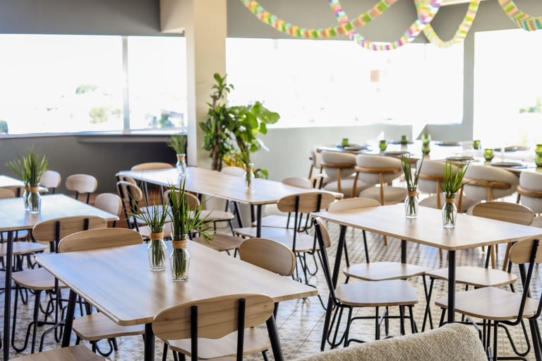 Bright modern dining space with wooden tables, beige chairs, potted plants, and festive yellow garland decorations overhead