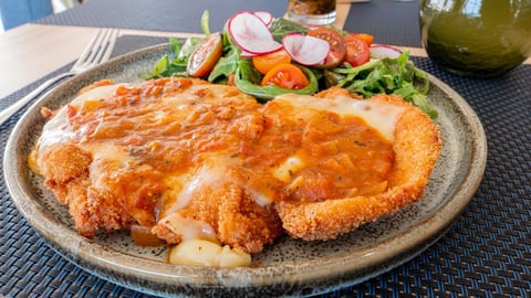 Crispy fried chicken cutlet topped with sauce, served with fresh salad and sliced radishes on a gray plate