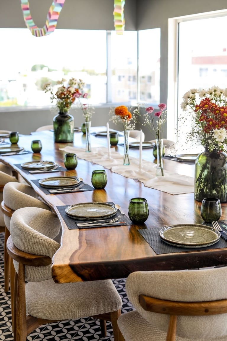 Elegant dining room table set with green glass vases, fresh flowers, place settings, and beige upholstered chairs along a wooden table