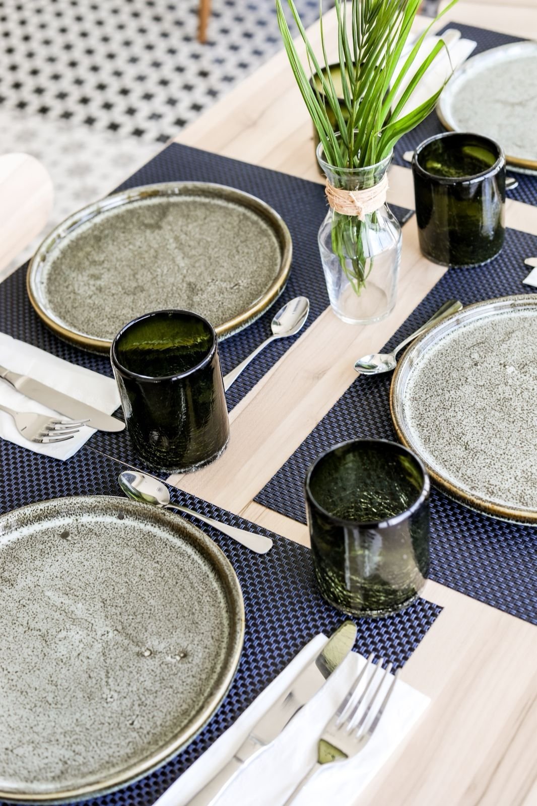 Elegant table setting with speckled plates, black cups, white napkins, and fresh green grass in a vase on navy and white runner