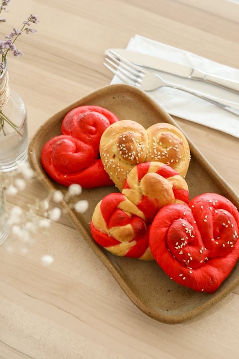 Heart-shaped pastries and sesame-topped buns arranged on a beige ceramic plate with white napkins and forks on a wooden table