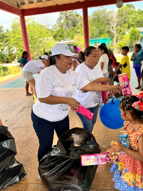 Volunteers in white shirts and caps distributing pink gift boxes to children at an outdoor community event under a red pavilion