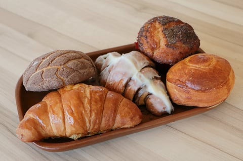 Assorted fresh baked breads and pastries in a wooden serving tray on a light wood surface