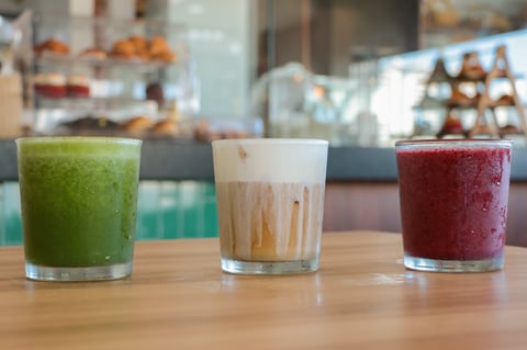 Three colorful beverages in glasses on a wooden counter: green juice on left, tan/caramel drink in center, and dark red juice on right